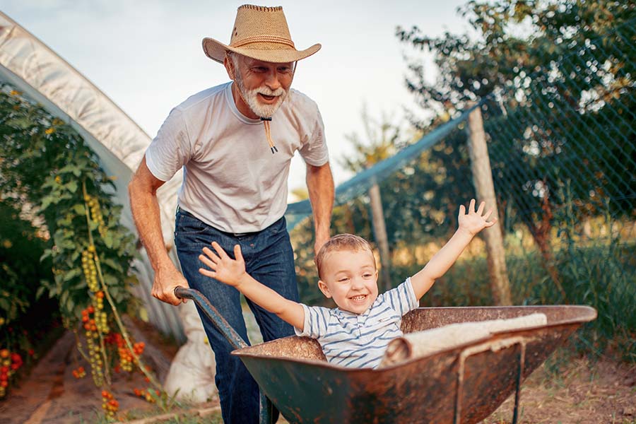 About Our Agency - View of Smiling Grandfather Pushing His Excited Grandson in a Wheelbarow in the Garden